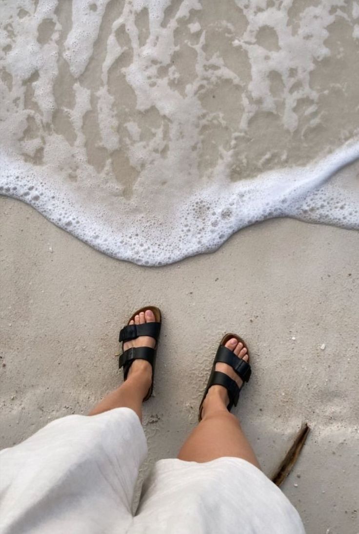 Black two-strap sandals on sandy beach with ocean foam, perfect for summer walks