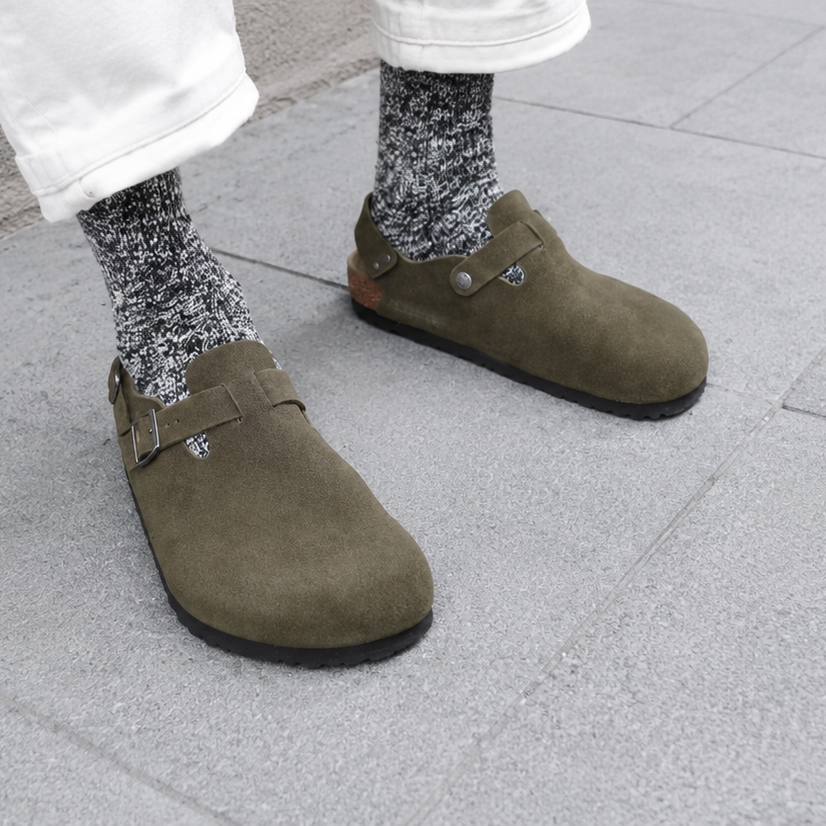 Olive green suede clogs with buckle straps, worn with patterned socks and white pants on pavement